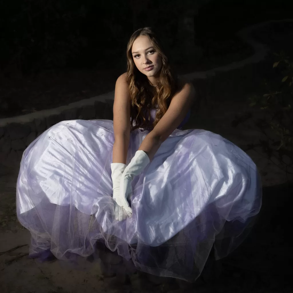 Jessica Welling sitting on a rock in a field of tall grass, wearing a flowing white dress.
