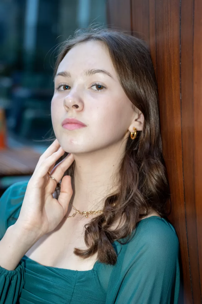 Lara, a teenage female, wearing a green top and gold jewelry, posing against a wooden background. The individual’s brown hair is styled in loose waves, 