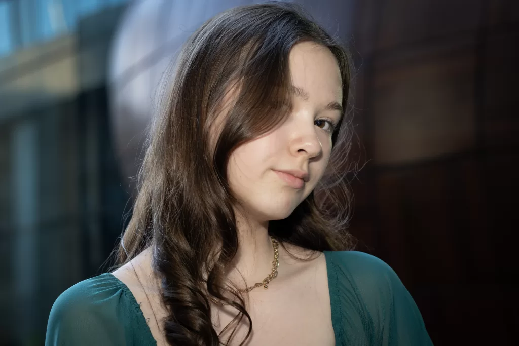 A teenage female wearing a green top and gold jewelry, The individual’s brown hair is styled in loose waves, 