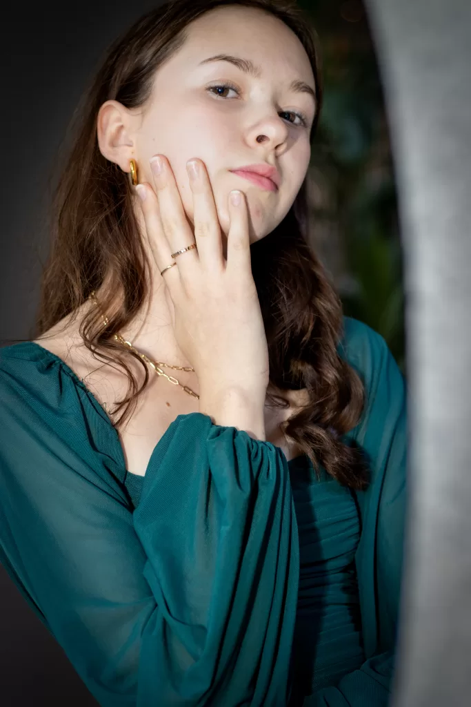 A teenage female wearing a green top and gold jewelry, posing against a wooden background. The individual’s brown hair is styled in loose waves, 