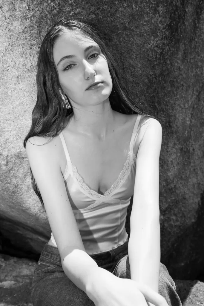 Black and white image of a teenage model wearng a silky top and posed delicately in an outdoor location.